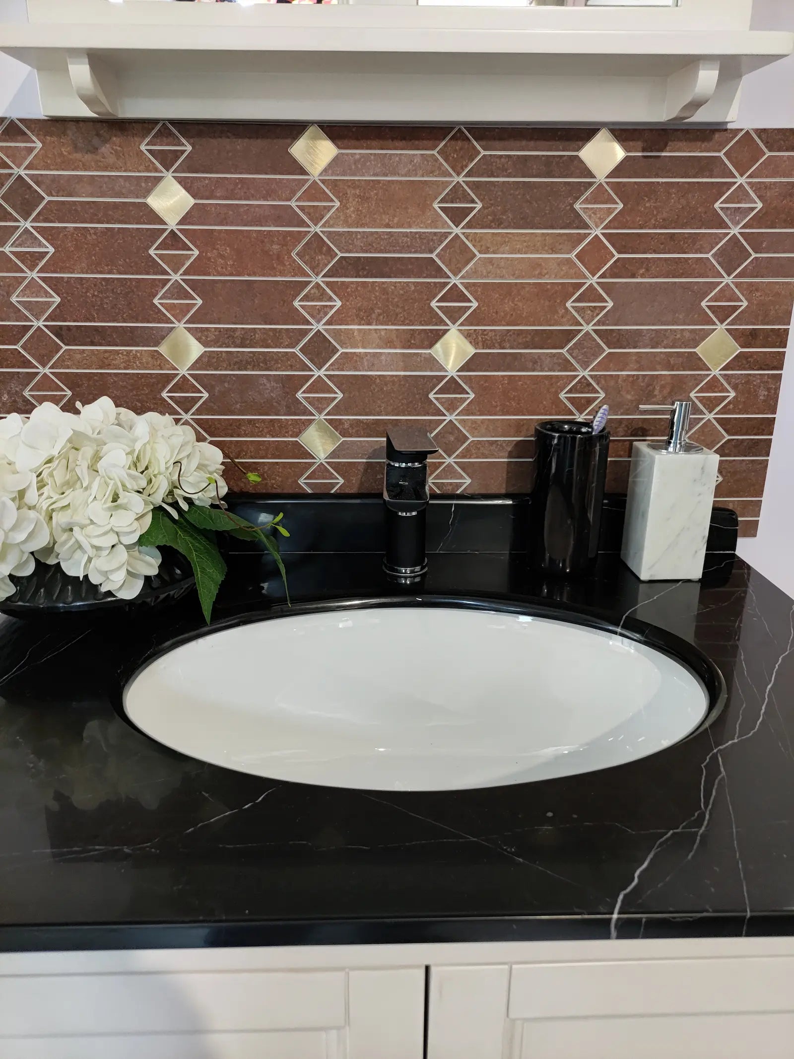 Full view of mid-century geometric peel and stick backsplash tile above a black countertop sink with decorative flowers and bathroom accessories